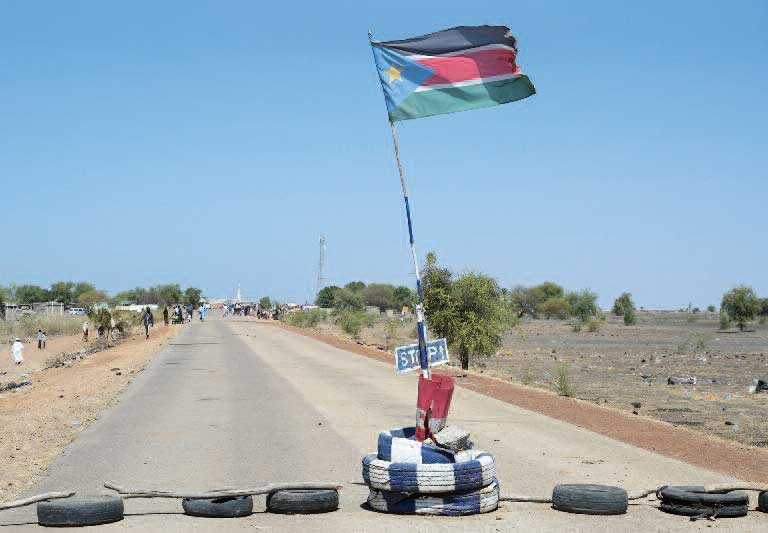 Mitten auf der Straße weht die Flagge des Südsudan. Darunter steht das Wort Stopp geschrieben. Der Durchgang ist mit alten Autoreifen und Ästen blockiert.