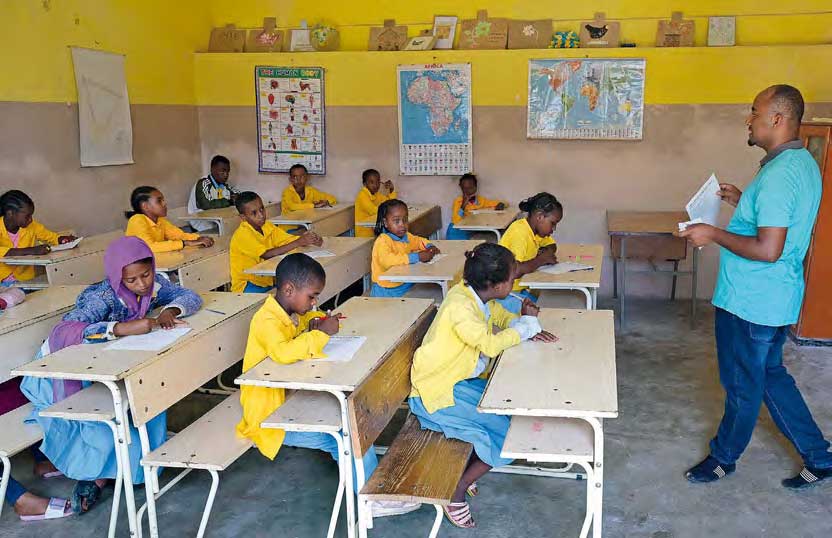Kinder sitzen in einem Klassenzimmer, jedes an einem eigenen Tisch. Ein Lehrer steht vorne und zeigt etwas