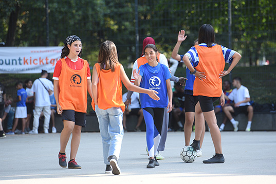 Mädchen in orangenen und blauen Trikots beim Handschlag nach dem Spiel auf einem Hartplatz. Eine von ihnen stellt den Fuß auf  einen Fußball.