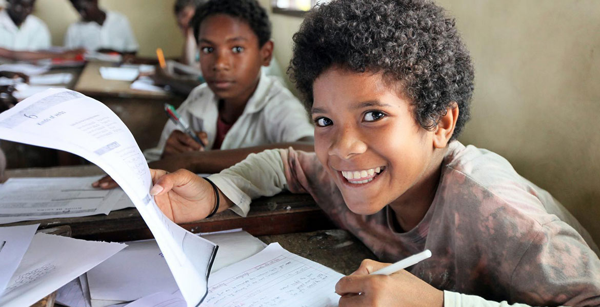 Ein Junge aus Papua-Neuguinea sitzt in der Schule und blickt lächelnd von den Unterlagen hoch.