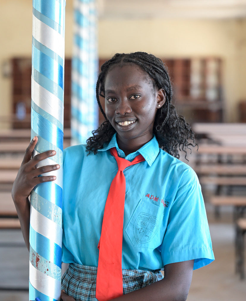 Ein heranwachsendes Mädchen, Cecilia A., steht in einem Klassenzimmer in Kakuma. Sie hat eine blaue Schuluniform an mit roter Krawatte und lächelt in die Kamera.