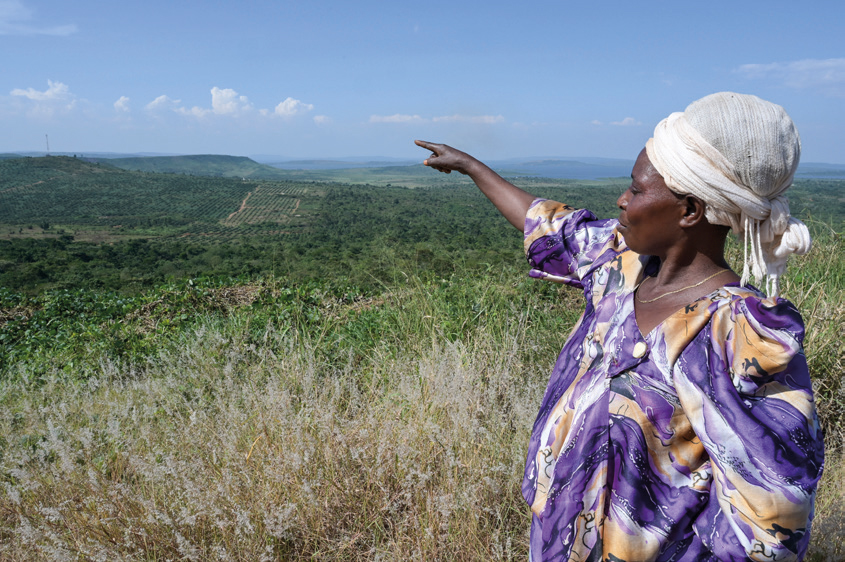Eine Frau mit beige-farbenem Kopftuch und lila-brauner Bluse steht auf einem Hügel in Uganda und zeigt in die Ferne, wo bewirtschaftete grüne Flächen zu sehen sind, soweit das Auge reicht.