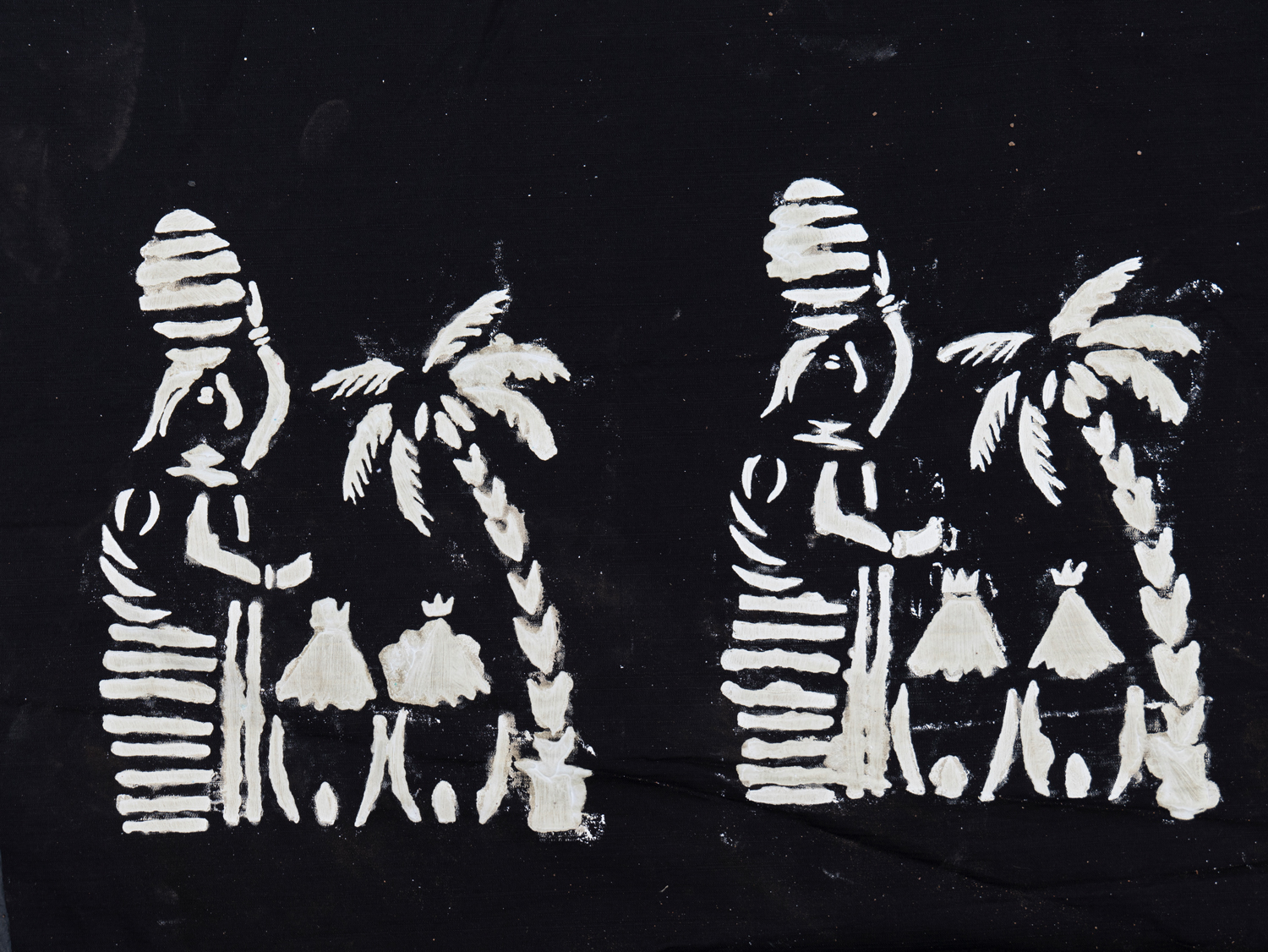 Ein weiß gebatiktes Motiv auf schwarzem Grund, das sich zweimal wiederholt. Es zeigt eine Frau, die ein Gefäß auf dem Kopf trägt, daneben zwei Hütten und eine Palme.