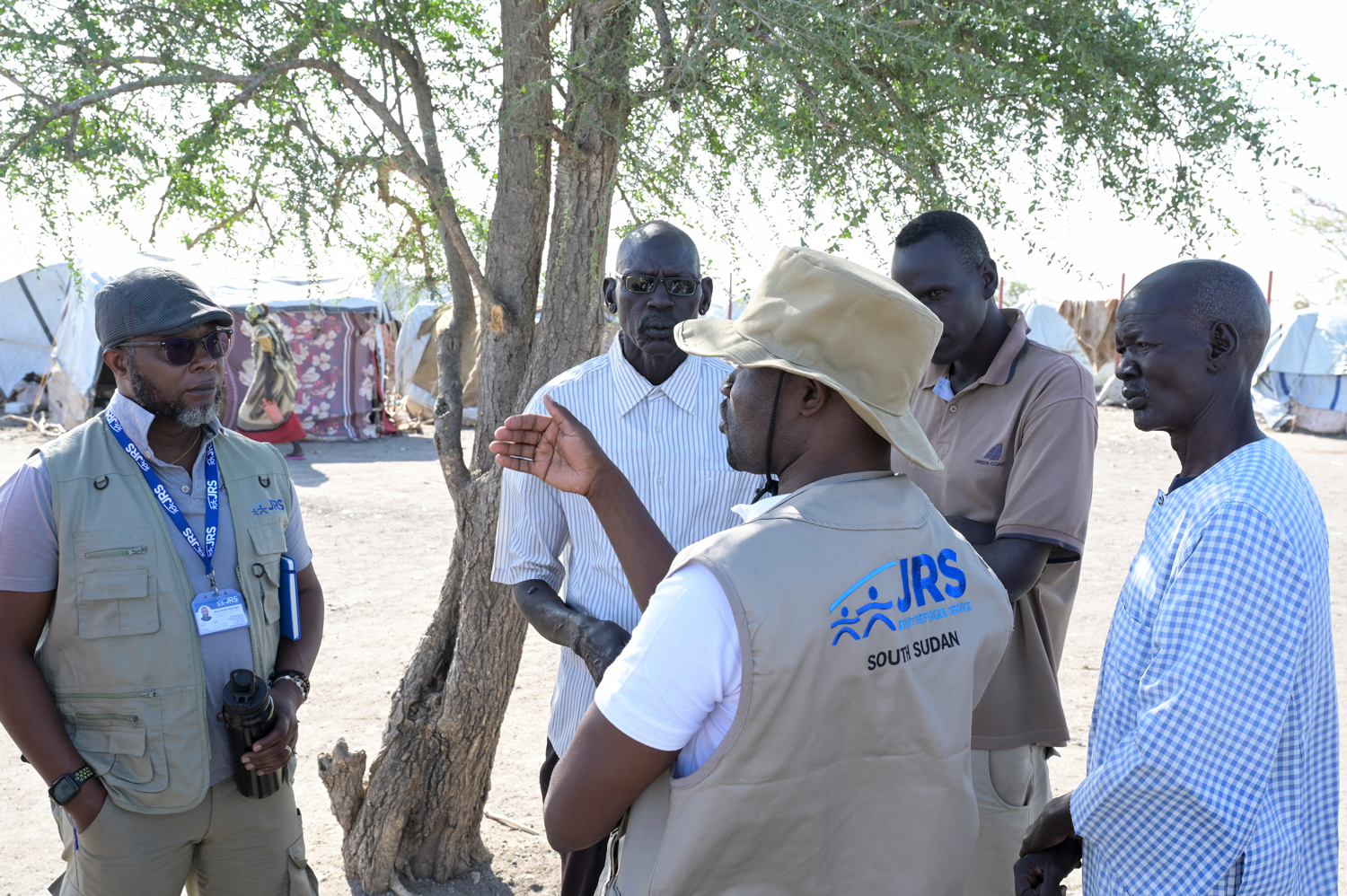 Regionaldirektor Andre Atsu und ein weiterer Mitarbeiter des Jesuitenflüchtlingsdienstes Südsudan sprechen mit drei Männern in mittlerem Alter auf einem Platz im Flüchtlingslager Renk.