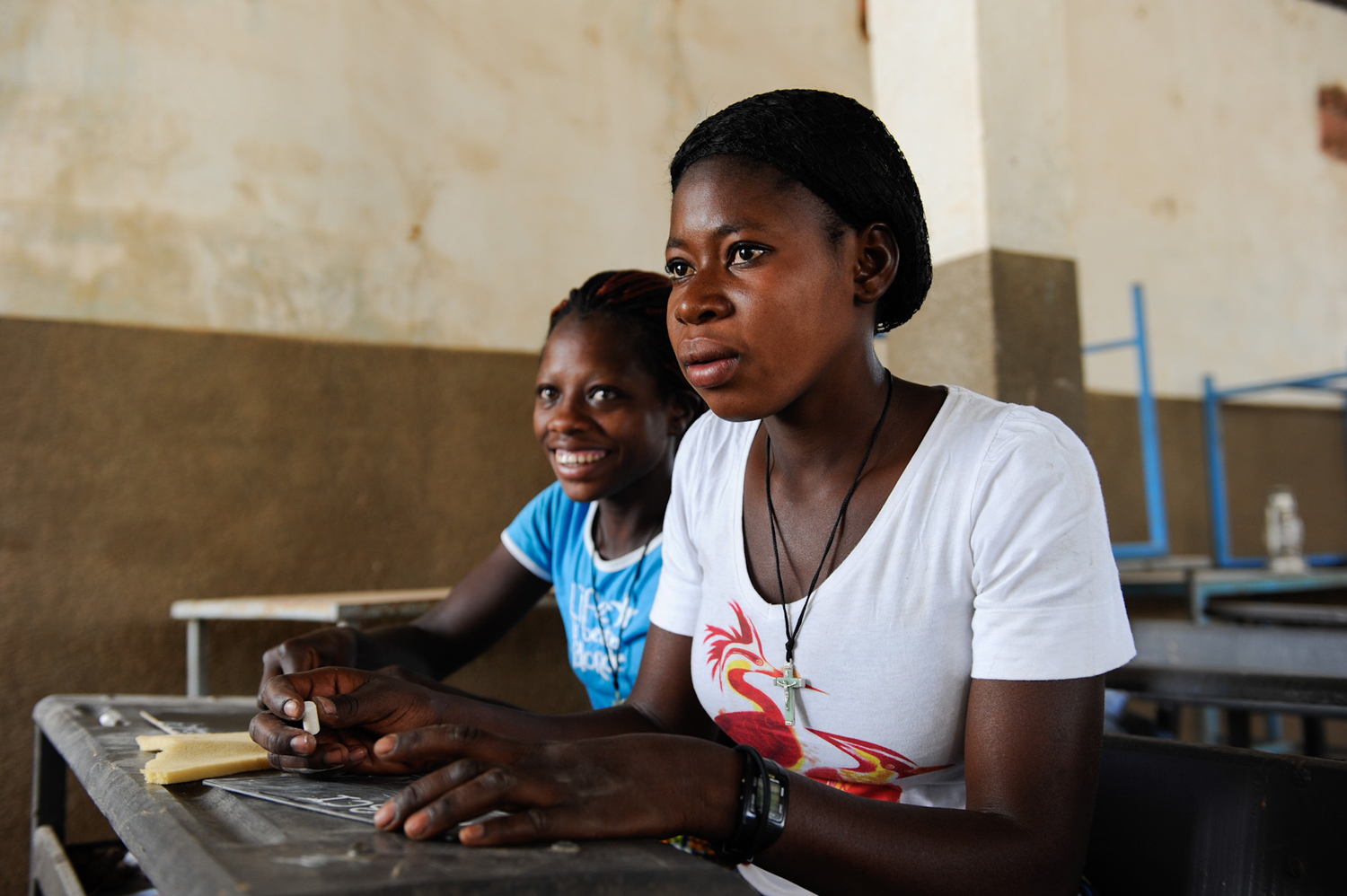 Zwei Mädchen aus Burkina Faso sitzen nebeneinander an einer Schulbank. Eine dreht den Kopf leicht und lächelt in die Kamera, die andere blickt konzentriert nach vorne.