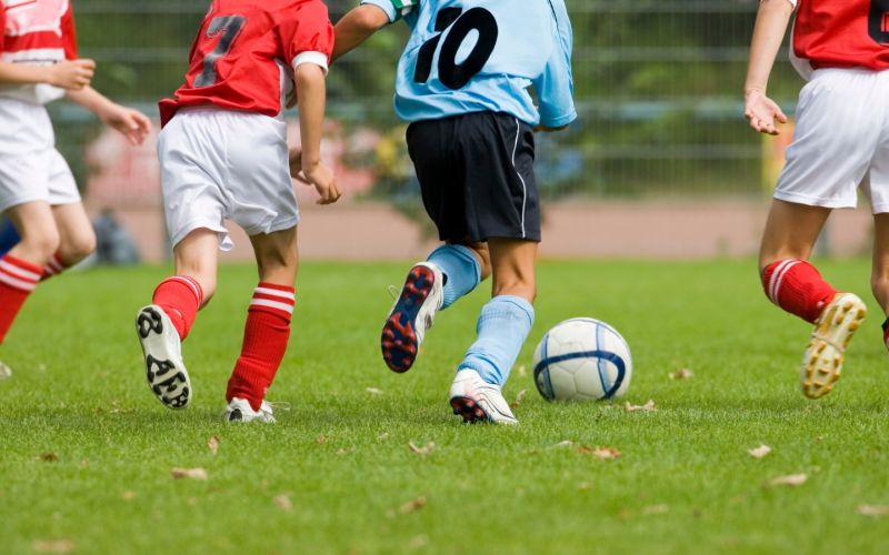 Auf einem Rasenplatz liegt ein Fußball. Vier Jungen bewegen sich auf den Ball zu. Zu sehen sind nur Beine und Rumpf. Drei Jungs tragen ein rotes Trikot mit weißer Hose, ein weiterer Junge ein blaues Trikot mit schwarzer Hose.