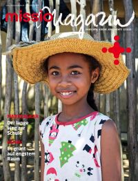 Titelbild missio magazin Ausgabe3/2026: Ein Mädchen aus Madagaskar  steht in ihrem Heimatdorf vor einem Zaun aus Holz. Sie trägt einen Sonnenhut und lacht in die Kamera.