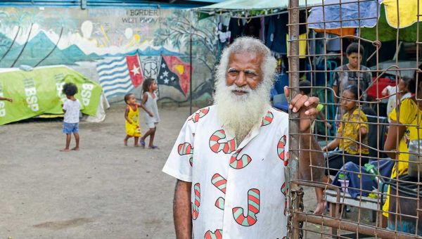 Ein Mann mit grauen Haaren und Bart steht am Tor eines Geflüchteten-Camps. Hinter ihm sind einige Kinder zu sehen. An einer Wand am Ende des Hofs sind zwei Flaggen gemalt, dahinter eine Inselwelt.