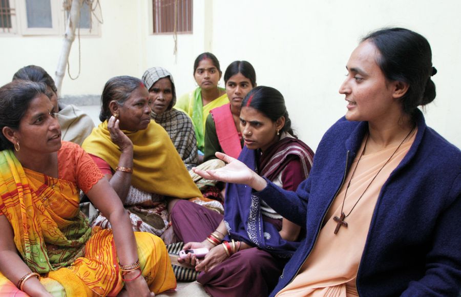 Eine junge Ordensschwester aus Indien spricht mit einer Frau in buntem Gewand, die ihr gegenüber auf dem Boden sitzt. Neben ihnen weitere Frauen unterschiedlichen Alters.