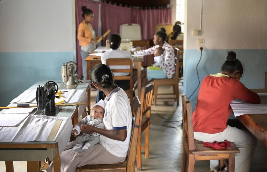 Frauen sitzen an Nähtischen und arbeiten. Eine von ihnen hat dabei ein Baby auf dem Schoß.
