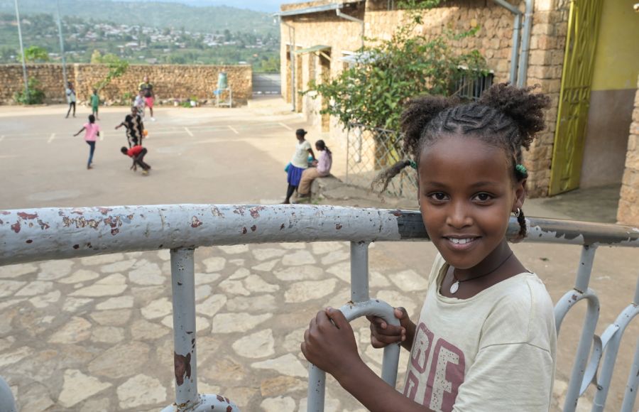 Ein Mädchen lächelt in die Kamera Sie hält sich an einem Geländer fest. Im Hintergrund sind Kinder in einem Innenhof zu sehen.