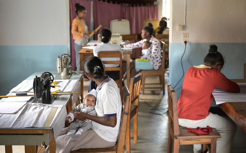 Frauen sitzen an Nähtischen und arbeiten. Eine von ihnen hat dabei ein Baby auf dem Schoß.