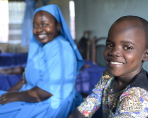 Ein Junge sitzt auf einem Bett im Kinderschutzzentrum Lusubilo in Karonga/Malawi und lacht in die Kamera. Im Hintergrund sitzt eine Ordensschwester und blickt ebenfalls lächelnd in die Kamera.