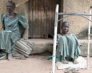 Zwei Frauen sitzen im Lepra-Dorf in Agok auf dem Boden. Eine hat vor sich eine Art Gehkonstruktion stehen.