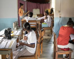 Frauen sitzen an Nähtischen und arbeiten. Eine von ihnen hat dabei ein Baby auf dem Schoß.