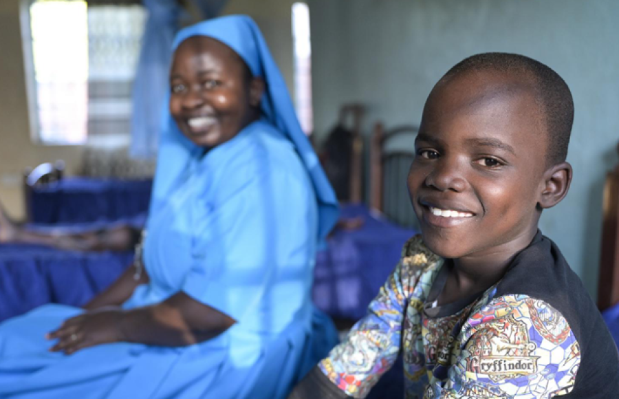 Ein Junge sitzt auf einem Bett im Kinderschutzzentrum Lusubilo in Karonga/Malawi und lacht in die Kamera. Im Hintergrund sitzt eine Ordensschwester und blickt ebenfalls lächelnd in die Kamera.