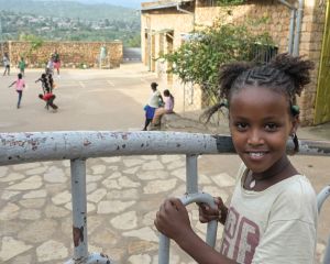 Ein Mädchen lächelt in die Kamera Sie hält sich an einem Geländer fest. Im Hintergrund sind Kinder in einem Innenhof zu sehen.